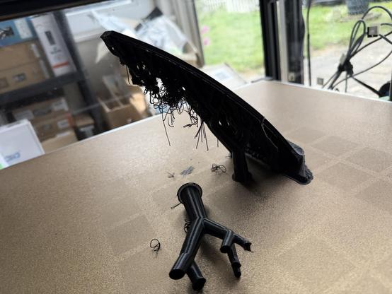 A black 3D-printed model with stringy strands and a branched base rests on a tan table near a window with boxes and grass outside.